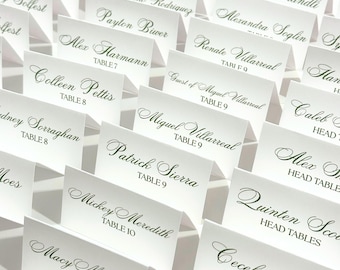 May include: A collection of white place cards with names and table numbers printed in elegant green script. The cards are arranged in rows, creating a formal and organized display, ideal for a wedding or event.