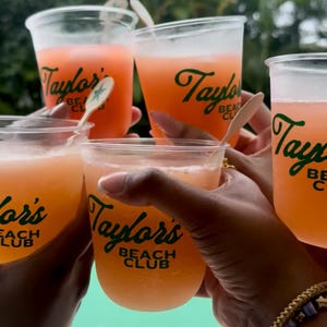 May include: Several hands holding up clear plastic cups filled with orange-colored drinks. The cups have the text "Taylor's Beach Club" in green script. Wooden spoons are in some of the drinks. The background is blurred, showing green foliage.