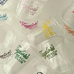 May include: Clear plastic cups with the text "Taylor's Beach Club" in various colors. The cups are stacked and scattered, showcasing the logo in green, purple, pink, yellow, and black.