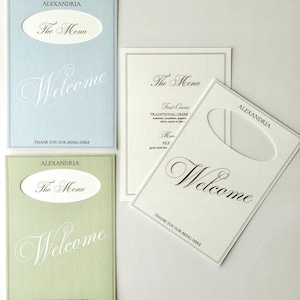May include: Three rectangular menu cards with die-cut oval windows. One is light blue, one is sage green, and one is cream. Each features the text Alexandria, The Menu, Welcome, and Thank You For Being Here in black script and serif fonts.