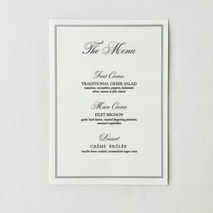 May include: A white rectangular menu card with a thin black double-line border. The text reads: The Menu, First Course: TRADITIONAL GREEK SALAD, Main Course: FILET MIGNON, and Dessert: CRÈME BRÛLÉE, along with descriptions of each dish in black ink.