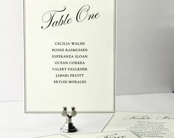Op de afbeelding: Een crèmekleurige tafelkaart voor een bruiloft met een zwarte dubbele lijnrand. De tekst luidt Table One, gevolgd door een lijst van zeven namen, waaronder Cecilia Walsh en Bodhi Rasmussen. De kaart wordt rechtop gehouden door een kleine zilveren metalen standaard.