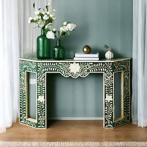 Può includere: Un tavolino da console verde con intarsi floreali bianchi intricati. Il tavolo ha un bordo a festoni ed è decorato con un motivo floreale. Il tavolo è su un pavimento in legno.