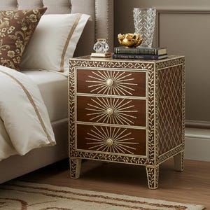 Może przedstawiać: Drewniana szafka nocna w kolorze ciemnobrązowym z trzema szufladami, z których każda ma wzór promieni słonecznych. Szafka ma kremowe akcenty i ozdobne wykończenia. W tle łóżko z białą pościelą i wzorzystą poduszką.
