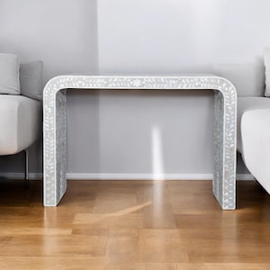 Puede incluir: Una mesa consola blanca con un diseño curvo e incrustaciones florales intrincadas. La mesa está hecha de madera y tiene un acabado blanco. La mesa está sobre un suelo de madera.