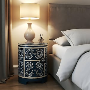 Op de afbeelding: Een blauwe en witte nachtkastje van beeninleg met twee laden en een bloemenmotief. Het nachtkastje staat naast een bed met witte beddengoed en een lamp met een witte kap.