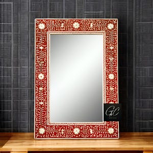 May include: A rectangular mirror with a decorative floral design. The mirror has a white frame with a red background and a white floral pattern. The mirror is on a wooden surface.