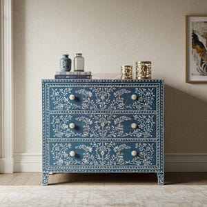 May include: A blue chest of drawers with three drawers, adorned with a white floral inlay design. The chest has round, off-white knobs and is set against a neutral wall. Decorative vases and candles sit on top.