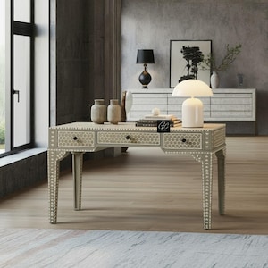 Puede incluir: Un escritorio de madera gris con tres cajones y detalles intrincados de incrustaciones de hueso. El escritorio tiene un patrón geométrico y se apoya en cuatro patas. El escritorio está en un ambiente de sala de estar moderna con una pared gris y una ventana en el fondo.