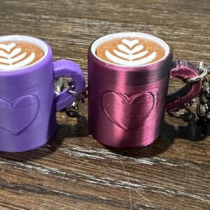 May include: Four small ceramic coffee mugs with a heart design and a latte art design on the top. The mugs are in different colors: teal, purple, burgundy, and gold.