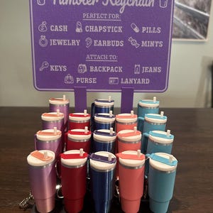 May include: A display of miniature tumbler keychains in various colors, including pink, red, blue, and teal. Each tumbler has a white lid and a straw. A purple sign above lists items the keychain can hold, such as cash, chapstick, and jewelry. The sign also lists items the keychain can attach to.
