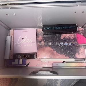 May include: An open silver makeup case filled with various cosmetic products. Items include eyeshadow palettes, a makeup sponge, lipstick, and other beauty supplies. The case is well-organized, showcasing a variety of colors and brands.