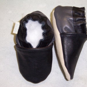 May include: A pair of black leather baby shoes with a gray leather trim. The shoes have a fold-over cuff and a soft, white lining.