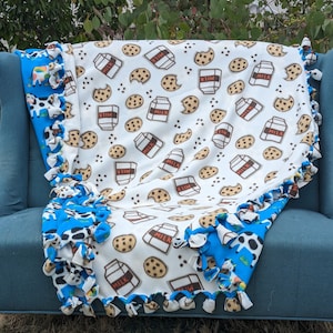 Pode incluir: Uma manta de lã branca com um padrão de caixa de leite e biscoitos. A manta é amarrada com tecido de estampa de vaca azul e preto.