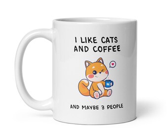 May include: A white ceramic mug featuring a cartoon orange cat holding a blue coffee cup. The text reads I LIKE CATS AND COFFEE AND MAYBE 3 PEOPLE. The cat has pink cheeks, a winking eye, and a small heart icon above its head. The design is centered.