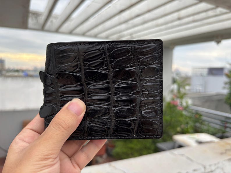 Puede incluir: Cartera de cuero negro con un patr&oacute;n de cocodrilo texturizado. La cartera es rectangular y tiene un cierre de solapa. La cartera est&aacute; sujeta por una mano.