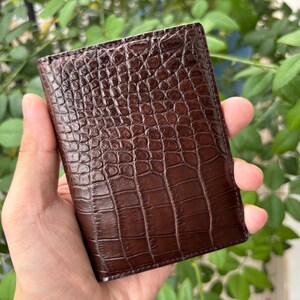 Pu&ograve; includere: Un portafoglio in pelle marrone con una trama a pelle di coccodrillo. Il portafoglio &egrave; rettangolare e sembra chiuso. Il portafoglio &egrave; tenuto in mano da una persona, con uno sfondo di foglie verdi.