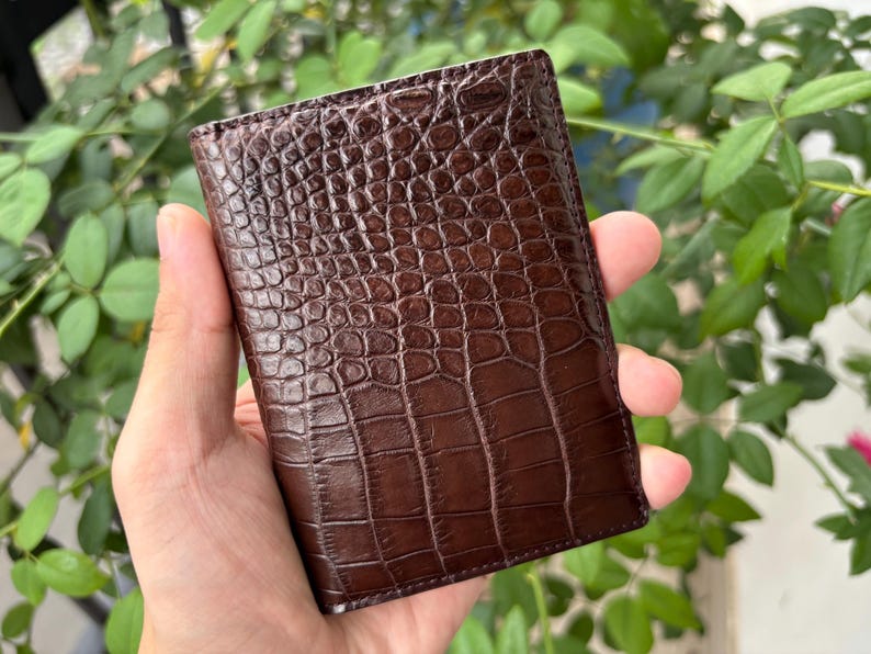 Pu&ograve; includere: Un portafoglio in pelle marrone con una trama a pelle di coccodrillo. Il portafoglio &egrave; rettangolare e sembra chiuso. Il portafoglio &egrave; tenuto in mano da una persona, con uno sfondo di foglie verdi.