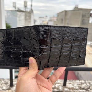 Puede incluir: Una cartera de cuero negro con un patr&oacute;n texturizado similar al de un cocodrilo. La cartera est&aacute; abierta, revelando el interior. El cuero tiene un acabado brillante. La cartera est&aacute; sujeta en la mano de una persona.