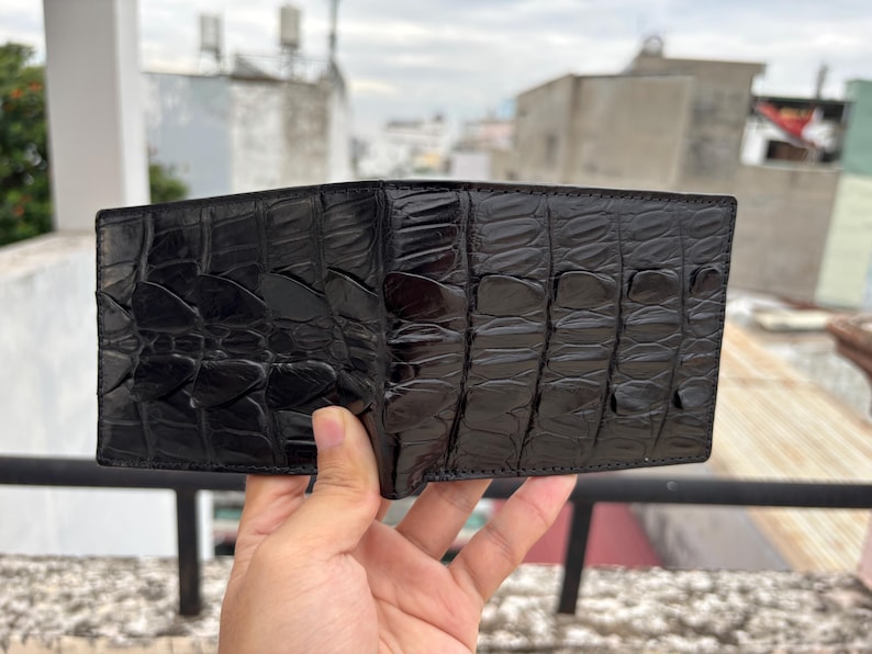 Puede incluir: Una cartera de cuero negro con un patr&oacute;n texturizado similar al de un cocodrilo. La cartera est&aacute; abierta, revelando el interior. El cuero tiene un acabado brillante. La cartera est&aacute; sujeta en la mano de una persona.