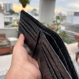 Puede incluir: Una cartera de cuero negro con un patr&oacute;n texturizado similar al de un reptil. La cartera est&aacute; abierta, revelando m&uacute;ltiples ranuras para tarjetas y compartimentos. La imagen est&aacute; tomada al aire libre con un fondo borroso de edificios y vegetaci&oacute;n.