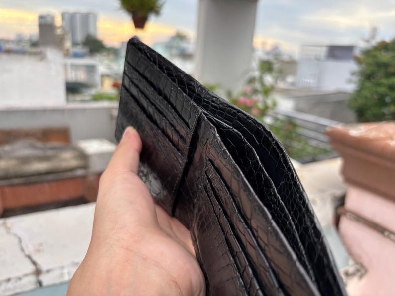 Puede incluir: Una cartera de cuero negro con un patr&oacute;n texturizado similar al de un reptil. La cartera est&aacute; abierta, revelando m&uacute;ltiples ranuras para tarjetas y compartimentos. La imagen est&aacute; tomada al aire libre con un fondo borroso de edificios y vegetaci&oacute;n.