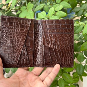Pu&ograve; includere: Portafoglio aperto in pelle marrone con una texture a pelle di coccodrillo. Il portafoglio ha pi&ugrave; fessure per carte e scomparti. Il portafoglio &egrave; tenuto aperto, mostrando il suo design interno. Lo sfondo presenta fogliame verde.