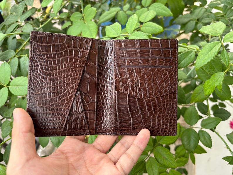Pu&ograve; includere: Portafoglio aperto in pelle marrone con una texture a pelle di coccodrillo. Il portafoglio ha pi&ugrave; fessure per carte e scomparti. Il portafoglio &egrave; tenuto aperto, mostrando il suo design interno. Lo sfondo presenta fogliame verde.
