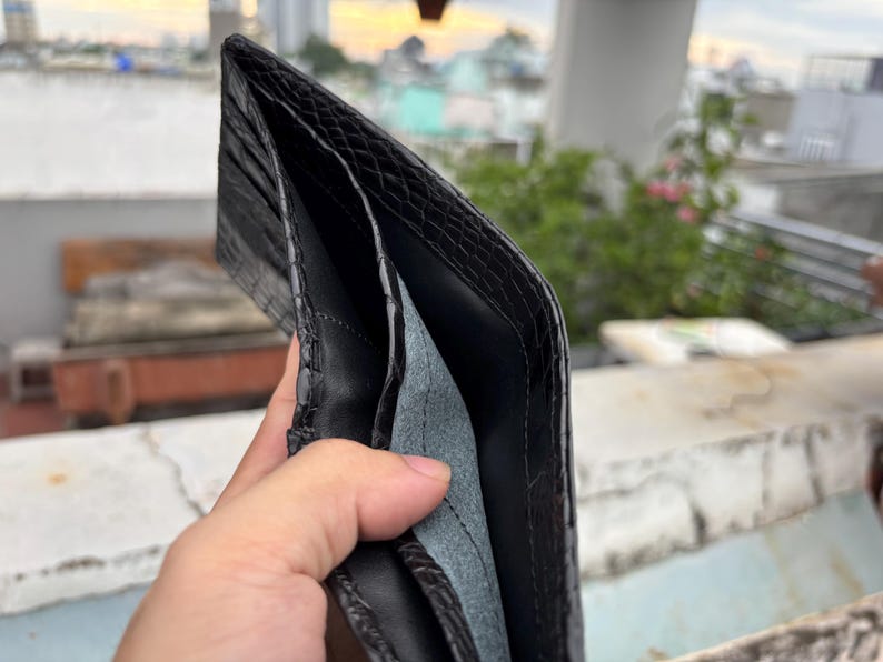 Puede incluir: Cartera de cuero negro abierta con un exterior texturizado. El interior presenta m&uacute;ltiples compartimentos y un forro gris claro. La cartera se sostiene abierta, mostrando su dise&ntilde;o y construcci&oacute;n.