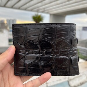 Puede incluir: Una cartera de cuero negro con un patr&oacute;n texturizado similar al de un cocodrilo. La cartera es rectangular y tiene un dise&ntilde;o de dos pliegues con un cierre de bot&oacute;n a presi&oacute;n. El cuero parece brillante.