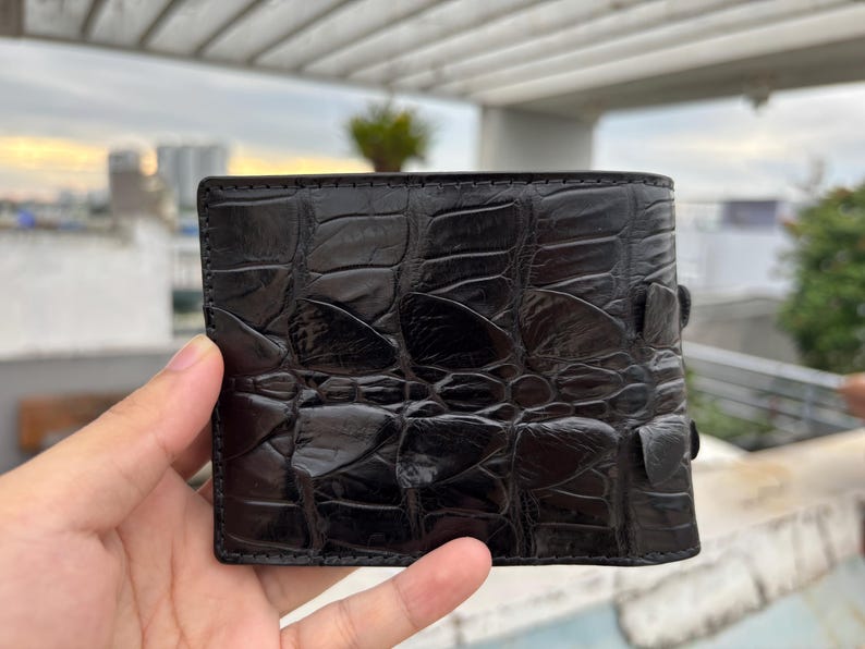 Puede incluir: Una cartera de cuero negro con un patr&oacute;n texturizado similar al de un cocodrilo. La cartera es rectangular y tiene un dise&ntilde;o de dos pliegues con un cierre de bot&oacute;n a presi&oacute;n. El cuero parece brillante.