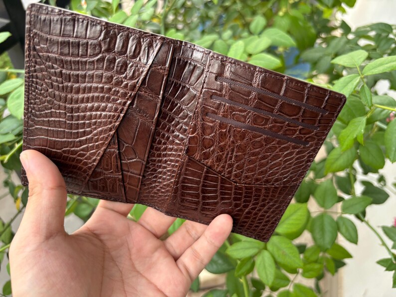 Pu&ograve; includere: Un portafoglio in pelle marrone con una texture a pelle di coccodrillo. Il portafoglio &egrave; aperto, rivelando pi&ugrave; fessure per carte e scomparti. La pelle ha un colore marrone scuro intenso e un motivo in rilievo dettagliato.