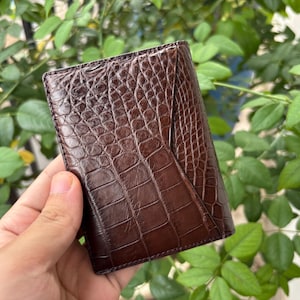 Pu&ograve; includere: Un portafoglio in pelle marrone con una texture a pelle di coccodrillo. Il portafoglio &egrave; rettangolare e ha un design piegato. Il colore &egrave; un marrone scuro intenso e la superficie presenta un motivo dettagliato a scaglie.