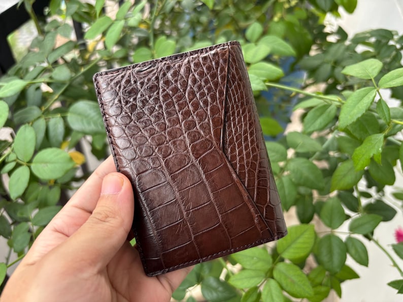 Pu&ograve; includere: Un portafoglio in pelle marrone con una texture a pelle di coccodrillo. Il portafoglio &egrave; rettangolare e ha un design piegato. Il colore &egrave; un marrone scuro intenso e la superficie presenta un motivo dettagliato a scaglie.