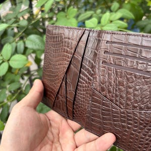 Pu&ograve; includere: Un portafoglio in pelle marrone con una trama a pelle di coccodrillo. Il portafoglio ha pi&ugrave; fessure e tasche per carte. Il portafoglio &egrave; tenuto aperto, rivelando il design interno. Lo sfondo &egrave; una vista sfocata di fogliame verde.