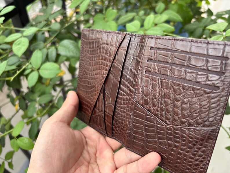 Pu&ograve; includere: Un portafoglio in pelle marrone con una trama a pelle di coccodrillo. Il portafoglio ha pi&ugrave; fessure e tasche per carte. Il portafoglio &egrave; tenuto aperto, rivelando il design interno. Lo sfondo &egrave; una vista sfocata di fogliame verde.