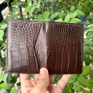 Pu&ograve; includere: Un portafoglio in pelle marrone con una trama a pelle di coccodrillo. Il portafoglio &egrave; rettangolare e ha una tasca sul lato sinistro. Il portafoglio &egrave; tenuto in mano da una persona. Lo sfondo &egrave; un fogliame verde sfocato.