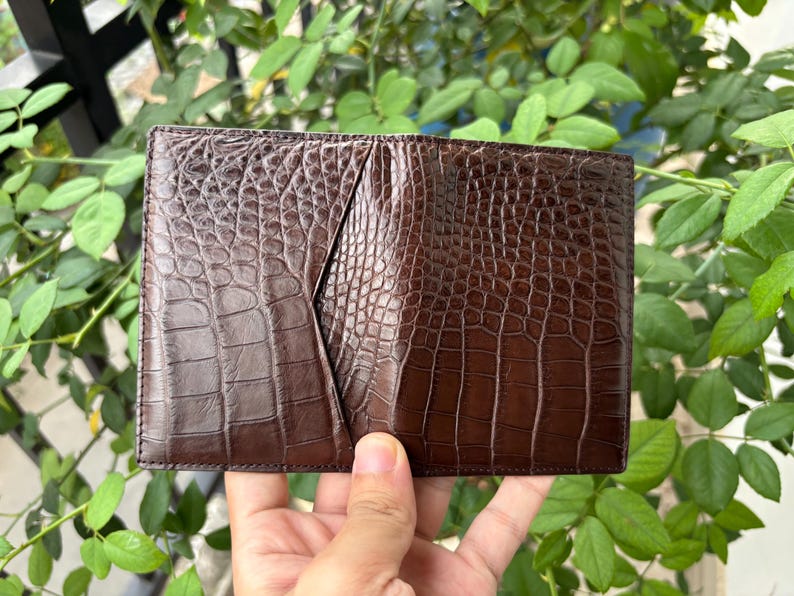 Pu&ograve; includere: Un portafoglio in pelle marrone con una trama a pelle di coccodrillo. Il portafoglio &egrave; rettangolare e ha una tasca sul lato sinistro. Il portafoglio &egrave; tenuto in mano da una persona. Lo sfondo &egrave; un fogliame verde sfocato.