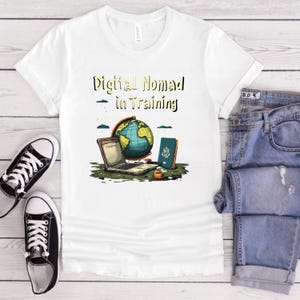 May include: White t-shirt with the text "Digital Nomad in Training" above a graphic of a laptop, globe, and passport. The shirt is paired with blue jeans and black and white sneakers.
