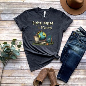 May include: Dark gray t-shirt with the text "Digital Nomad in Training" and a graphic of a laptop, globe, and passport. The shirt is paired with blue jeans, brown shoes, and a brown hat on a wooden surface.