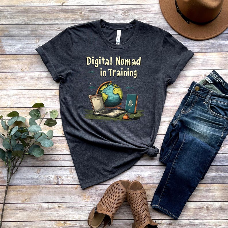 May include: Dark gray t-shirt with the text "Digital Nomad in Training" and a graphic of a laptop, globe, and passport. The shirt is paired with blue jeans, brown shoes, and a brown hat on a wooden surface.