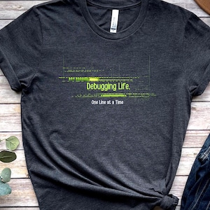 Könnte beinhalten: Dunkelgraues T-Shirt mit dem grünen Text "Debugging Life. One Line at a Time". Das Shirt hat einen Rundhalsausschnitt und kurze Ärmel. Der Text ist in einer modernen, serifenlosen Schriftart.