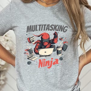 Pode incluir: Camiseta cinza com um gráfico de um ninja vermelho fazendo várias tarefas ao mesmo tempo com um laptop, café e um telefone. O texto "Multitasking Ninja" está impresso na camiseta.