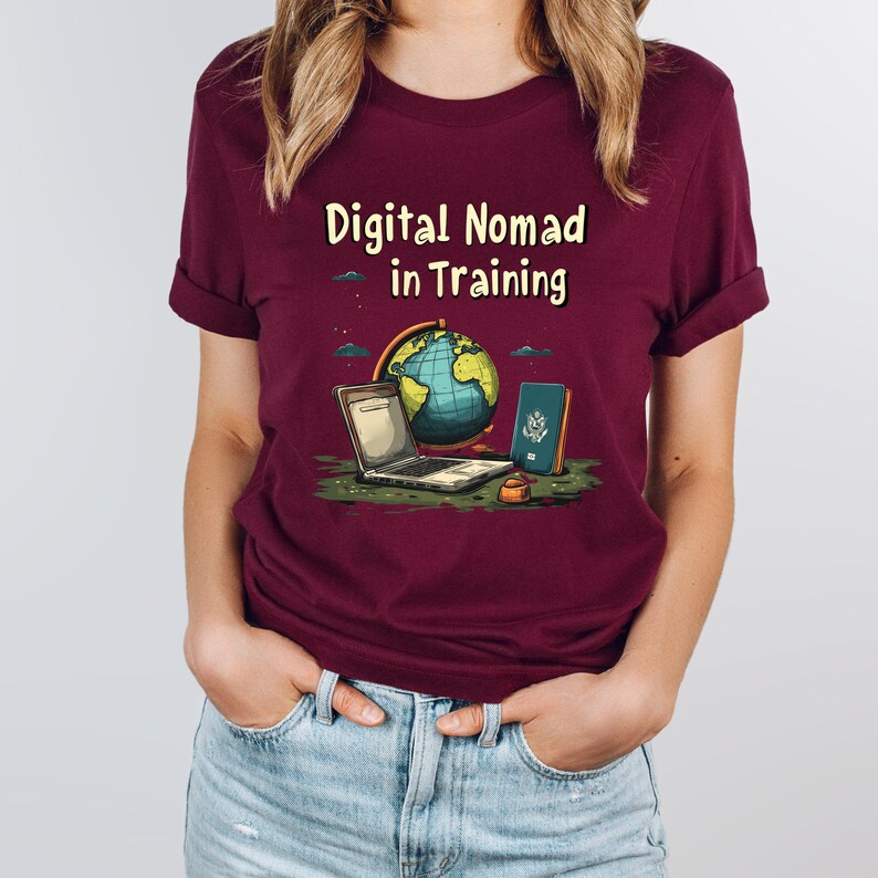 May include: A maroon t-shirt with rolled sleeves featuring the text "Digital Nomad in Training." The graphic includes a laptop, globe, passport, and a small bowl. The shirt is worn by a person in blue jeans.