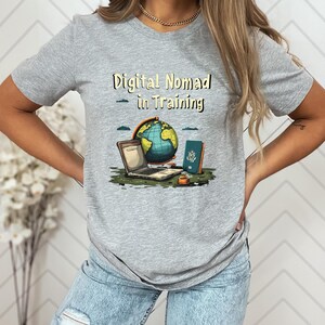 May include: A gray t-shirt with the text "Digital Nomad in Training" above a graphic of a laptop, globe, passport, and coffee cup. The globe is blue and green, and the passport is blue.