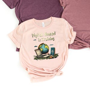 May include: A light pink t-shirt with the text "Digital Nomad in Training" above a graphic of a globe, laptop, passport, and other items. The shirt is made of soft material and has short sleeves. The background includes other t-shirts.