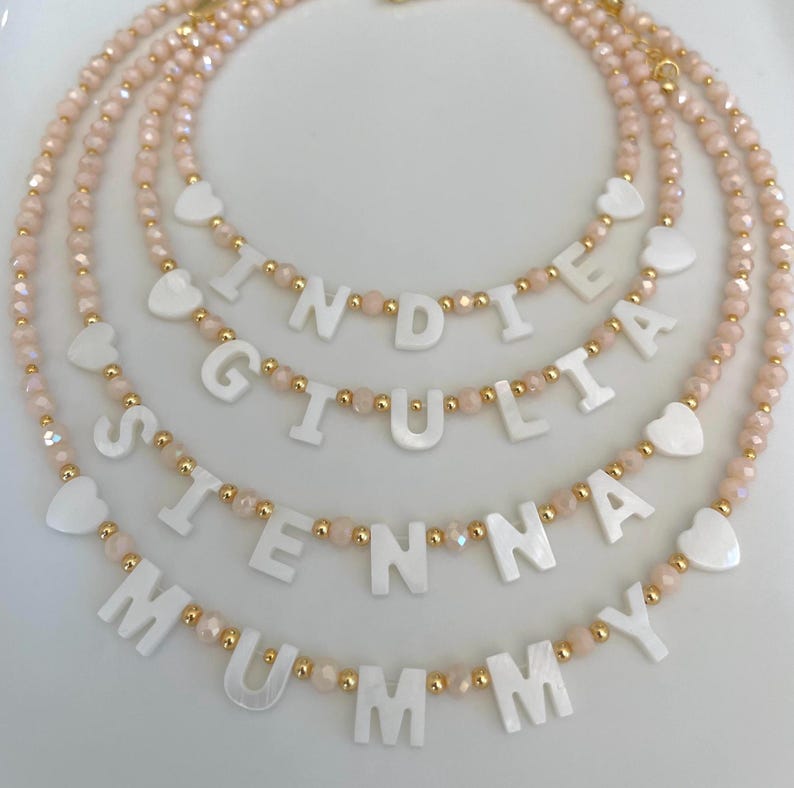 May include: Four layered necklaces featuring pale pink faceted beads and small gold-tone spacer beads. Each necklace includes white mother-of-pearl letter beads spelling out the names INDIE, GIULIA, SIENNA, and MUMMY, accented with white heart beads.