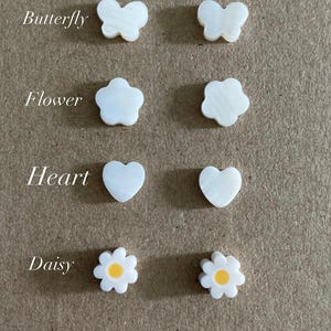 May include: A collection of small charms labeled Pick your charm, featuring pairs of white butterfly, flower, heart, and star shapes, plus a pair of white daisy charms with yellow centers. Each charm has a smooth, pearlescent finish and flat surface.