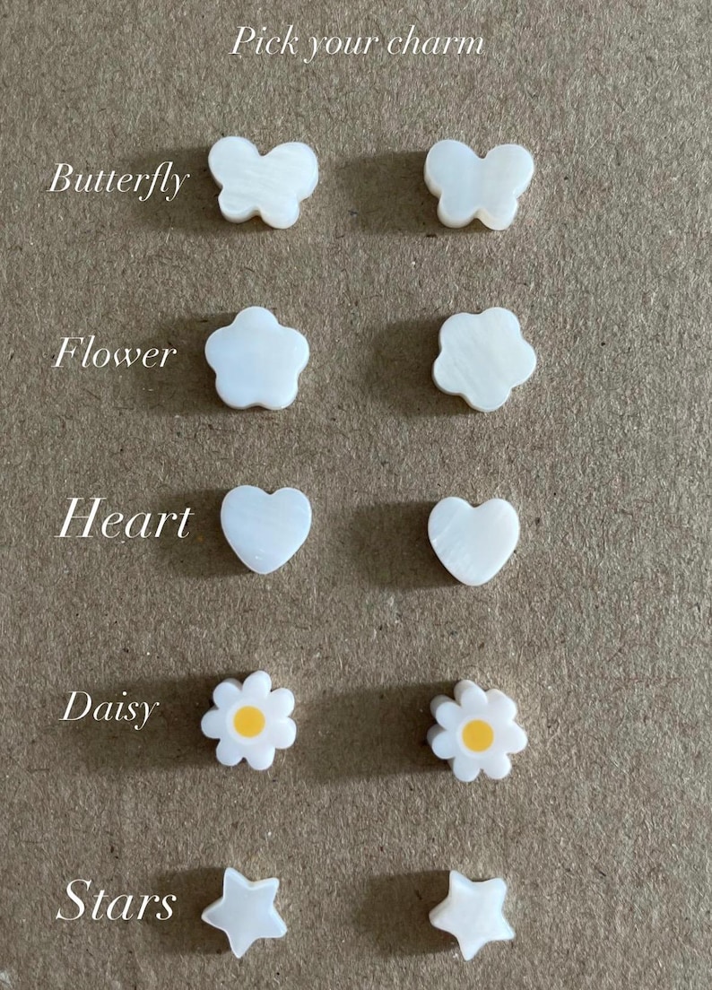 May include: A collection of small charms labeled Pick your charm, featuring pairs of white butterfly, flower, heart, and star shapes, plus a pair of white daisy charms with yellow centers. Each charm has a smooth, pearlescent finish and flat surface.
