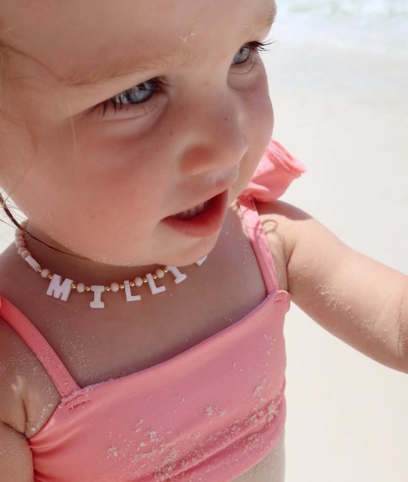 May include: A beaded necklace featuring white letter beads spelling the name MILLIE, interspersed with small gold-toned metallic beads and pale pink cylindrical beads, worn around the neck against a plain pink garment.